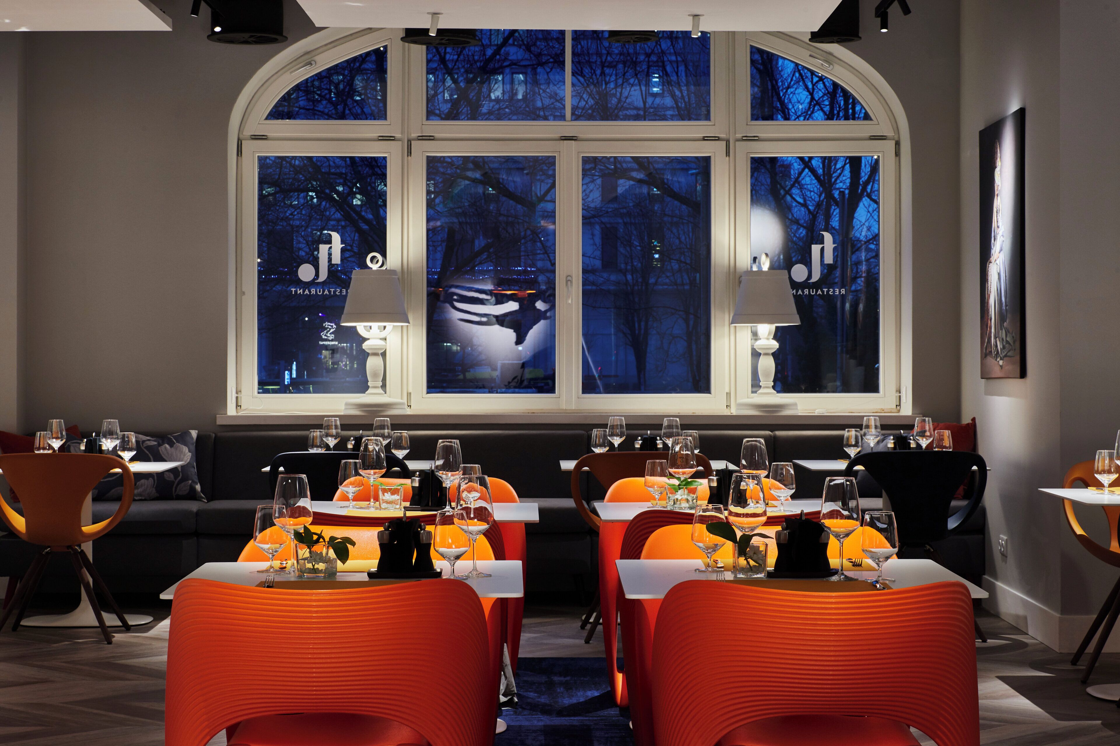 A restaurant with tables set for dining, a large arched window with a view of trees, and a modern interior design.
