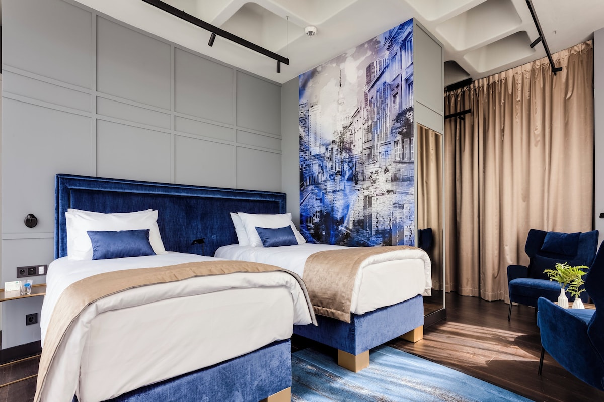 Ein modernes Hotelzimmer mit zwei Betten, einem Stadtbild-Wandgemälde, einem blauen Sessel und einem großen Fenster mit Vorhängen.
