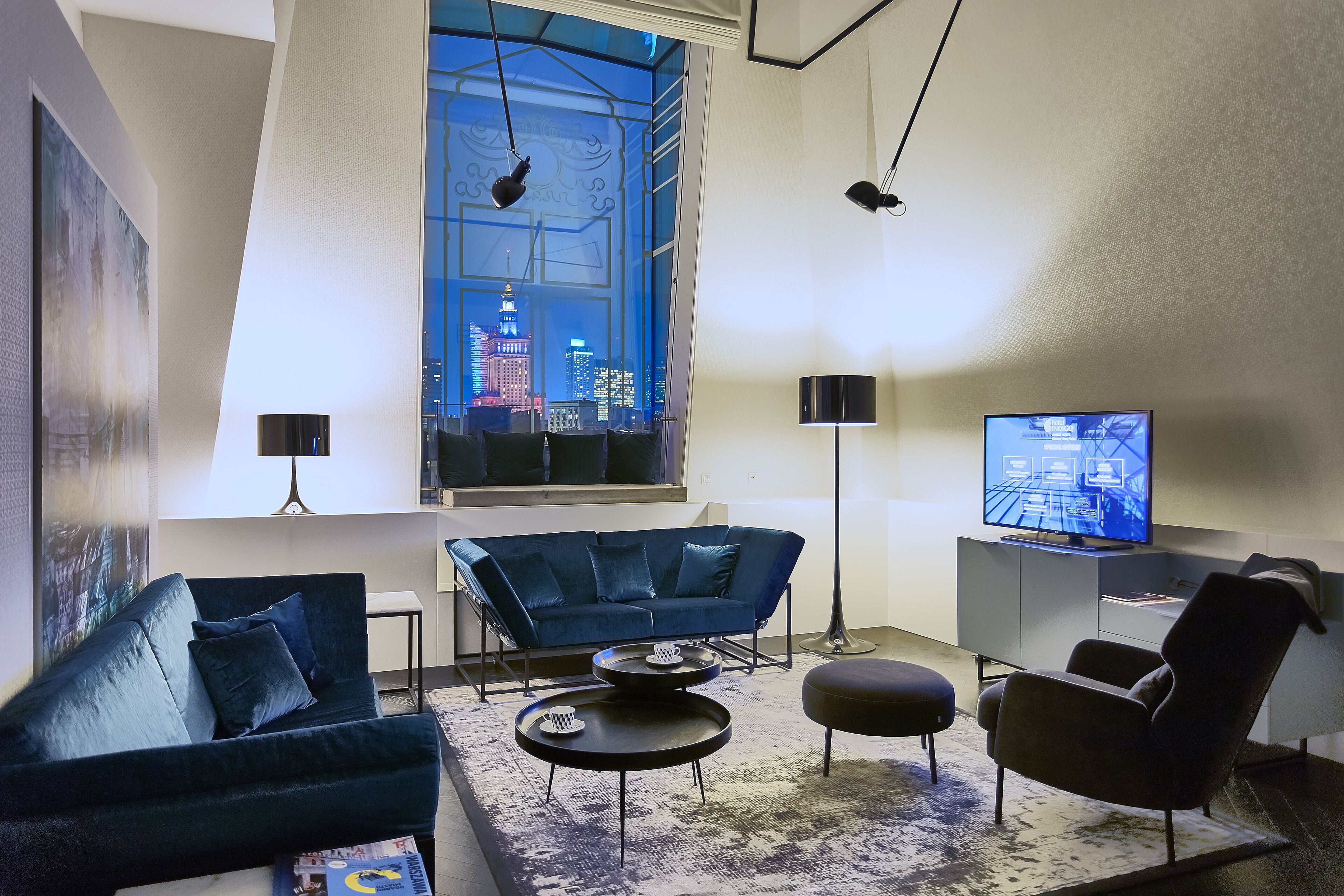 A modern living room with a dark blue sofa, a coffee table, and a TV. A large window offers a view of a cityscape at night.