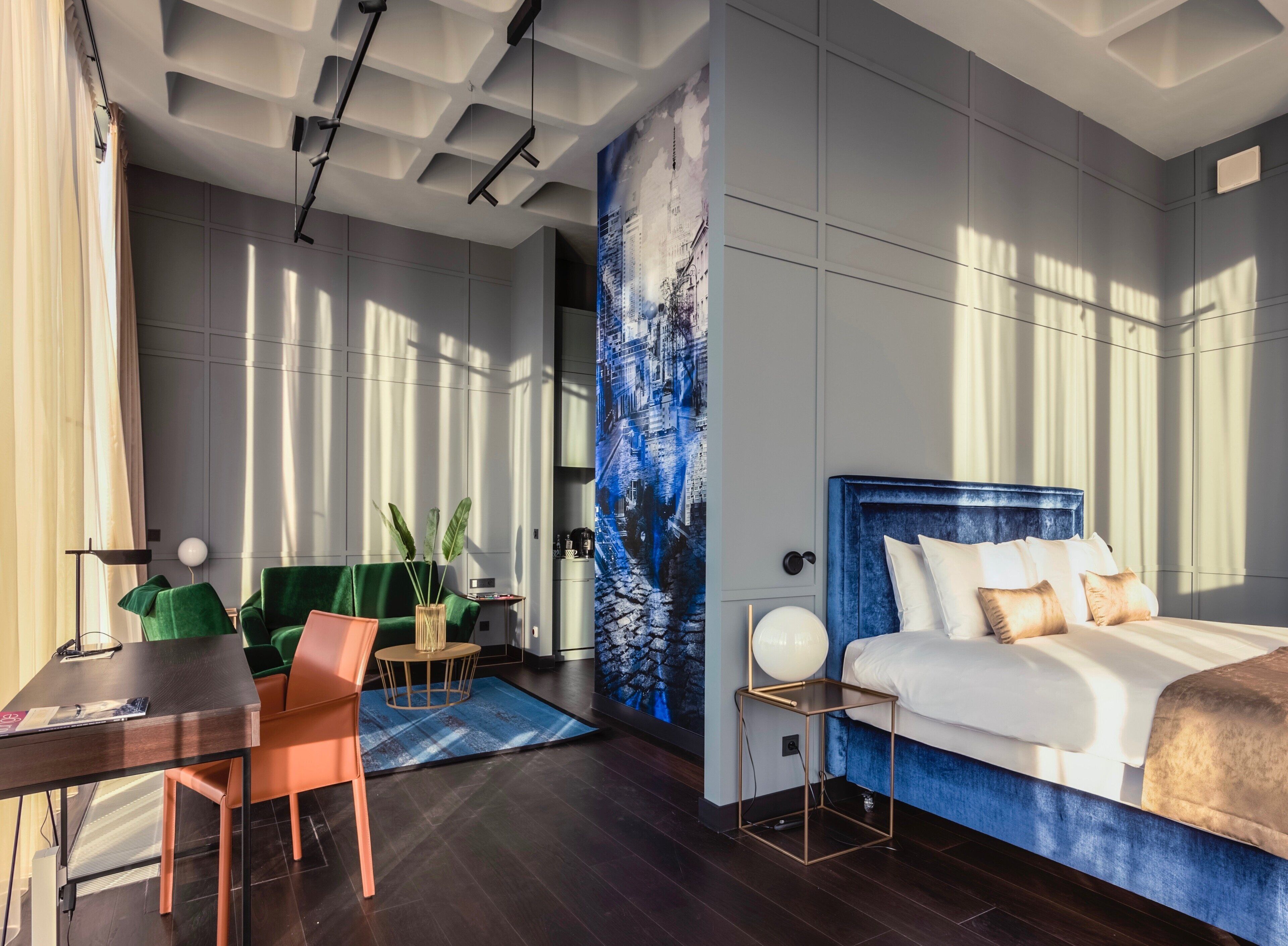 A modern hotel room with a bed, a desk, a chair, a small table with a plant, and a large window with natural light.