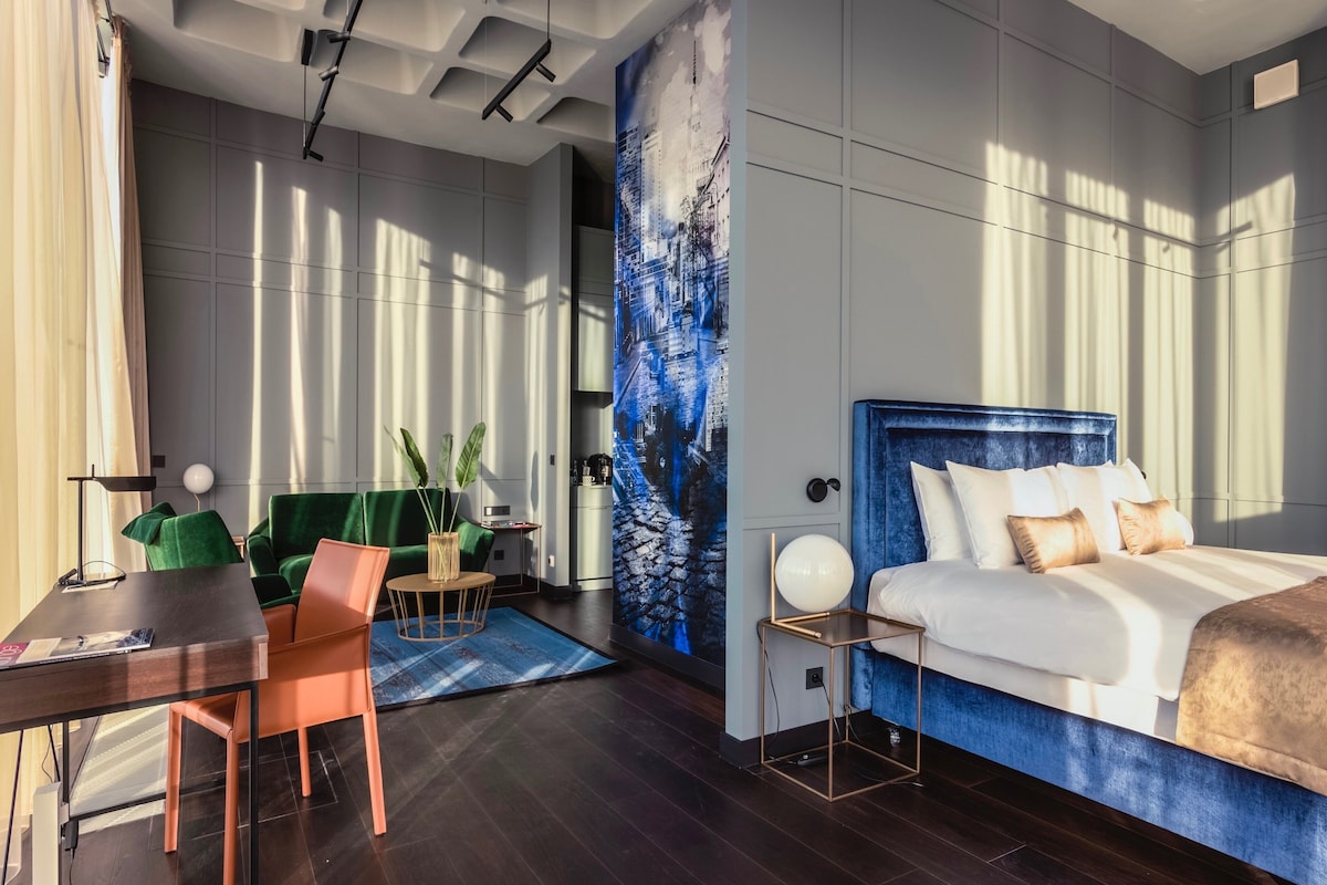 Ein modernes Hotelzimmer mit einem Bett, einem Schreibtisch, einem Stuhl, einem kleinen Tisch mit Pflanze und einem großen Fenster mit Tageslicht.
