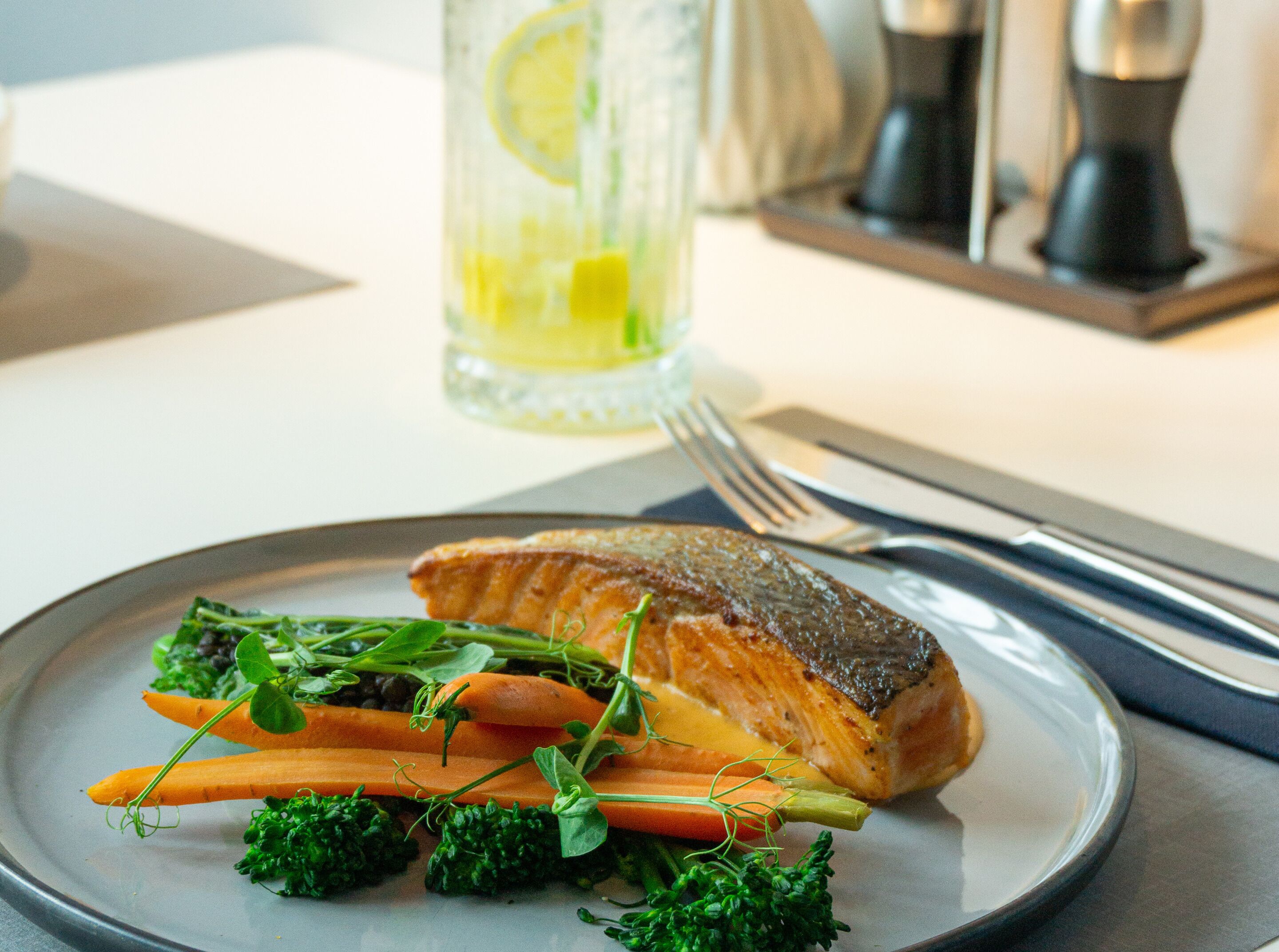 A plate with salmon, carrots, and greens, a glass of lemon water, and a fork and knife on a table.