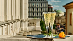 Deux flûtes à champagne sont posées sur un plateau, avec une fleur à côté, le tout sur fond d’une vue urbaine caractérisée par une architecture classique.