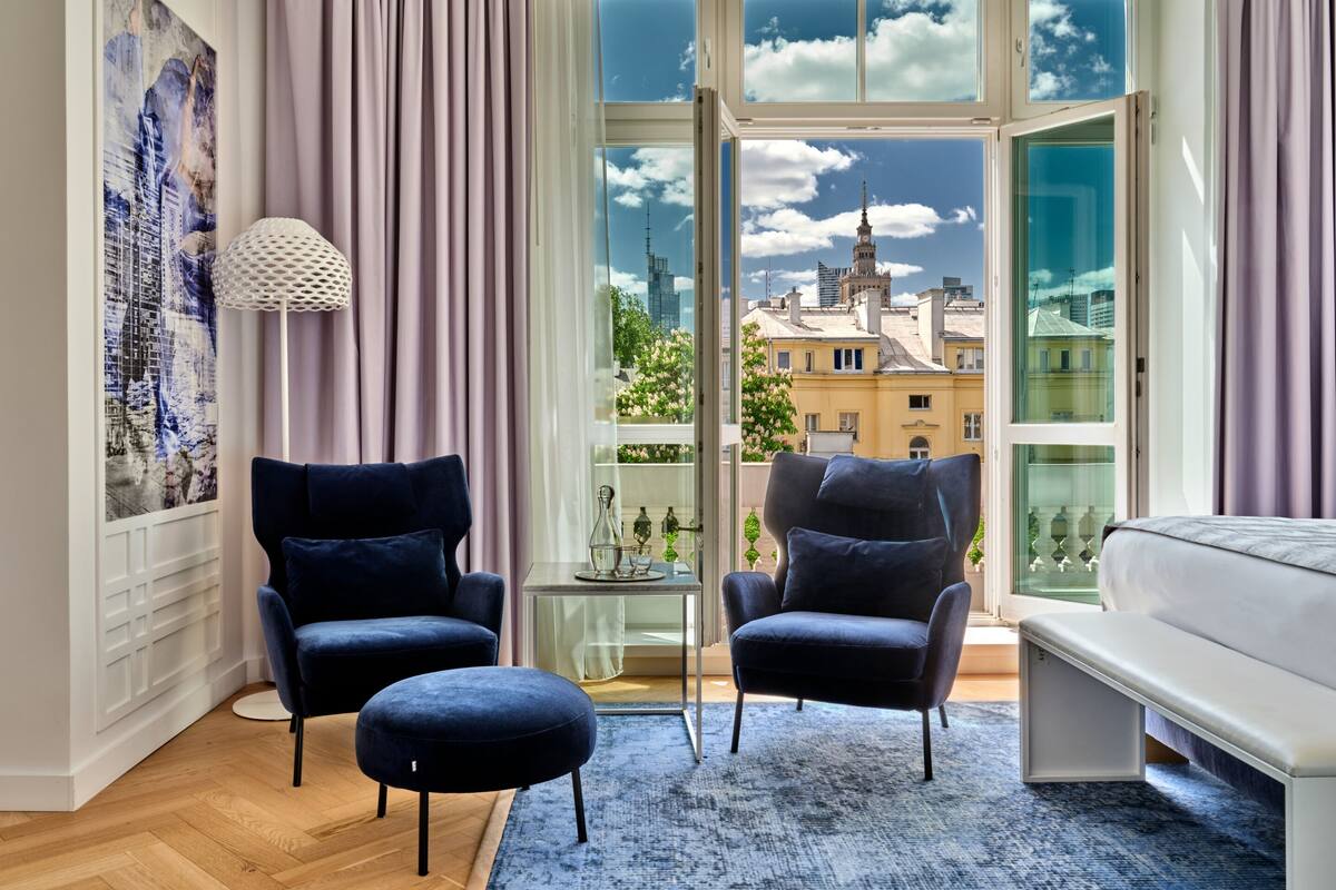 Een moderne slaapkamer met een groot raam, twee donkere fauteuils, een wit bed en een blauw vloerkleed.