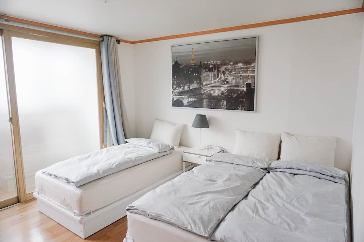 A double bed with a nightstand and a lamp, a window with curtains, and a cityscape painting on the wall.