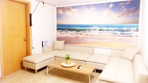 A room with a large beach mural, a wooden door, a beige sofa, a small table with a plant, and a sign that reads "Hong Kong One House." - Hongdae One House (Seoul)