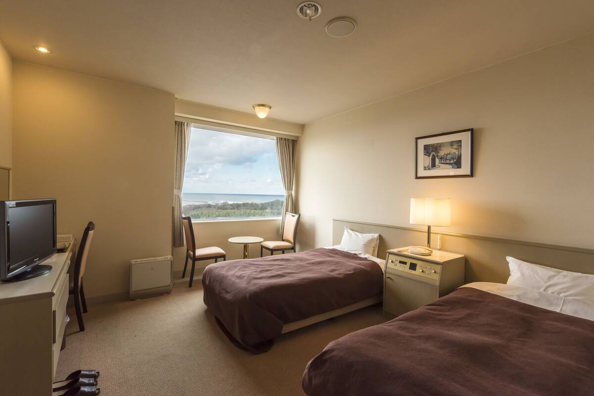A hotel room with a bed, a desk with a computer, a chair, a small table, and a window with a view.