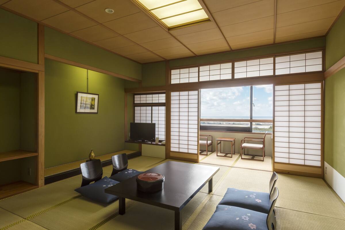 A traditional Japanese room with tatami flooring, a low table with cushions, a television, and large shoji screens.