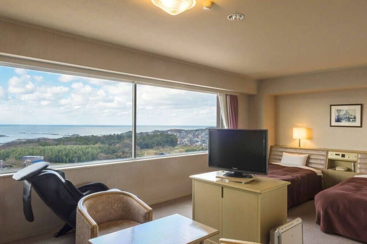A hotel room with a large window, a television, two beds, a desk, and a chair.