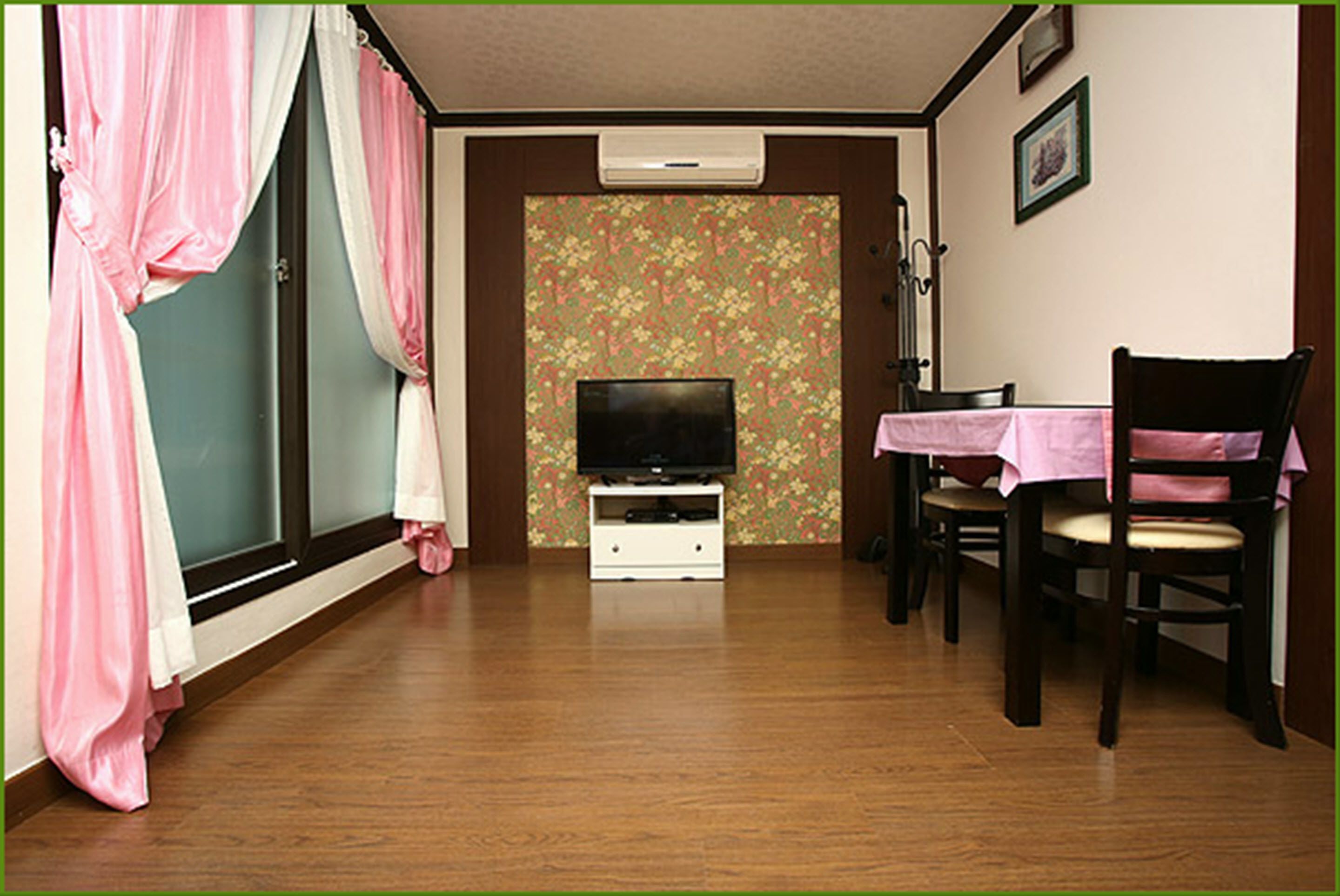 A room with a television on a white stand, a dining table with a pink tablecloth, and a chair.