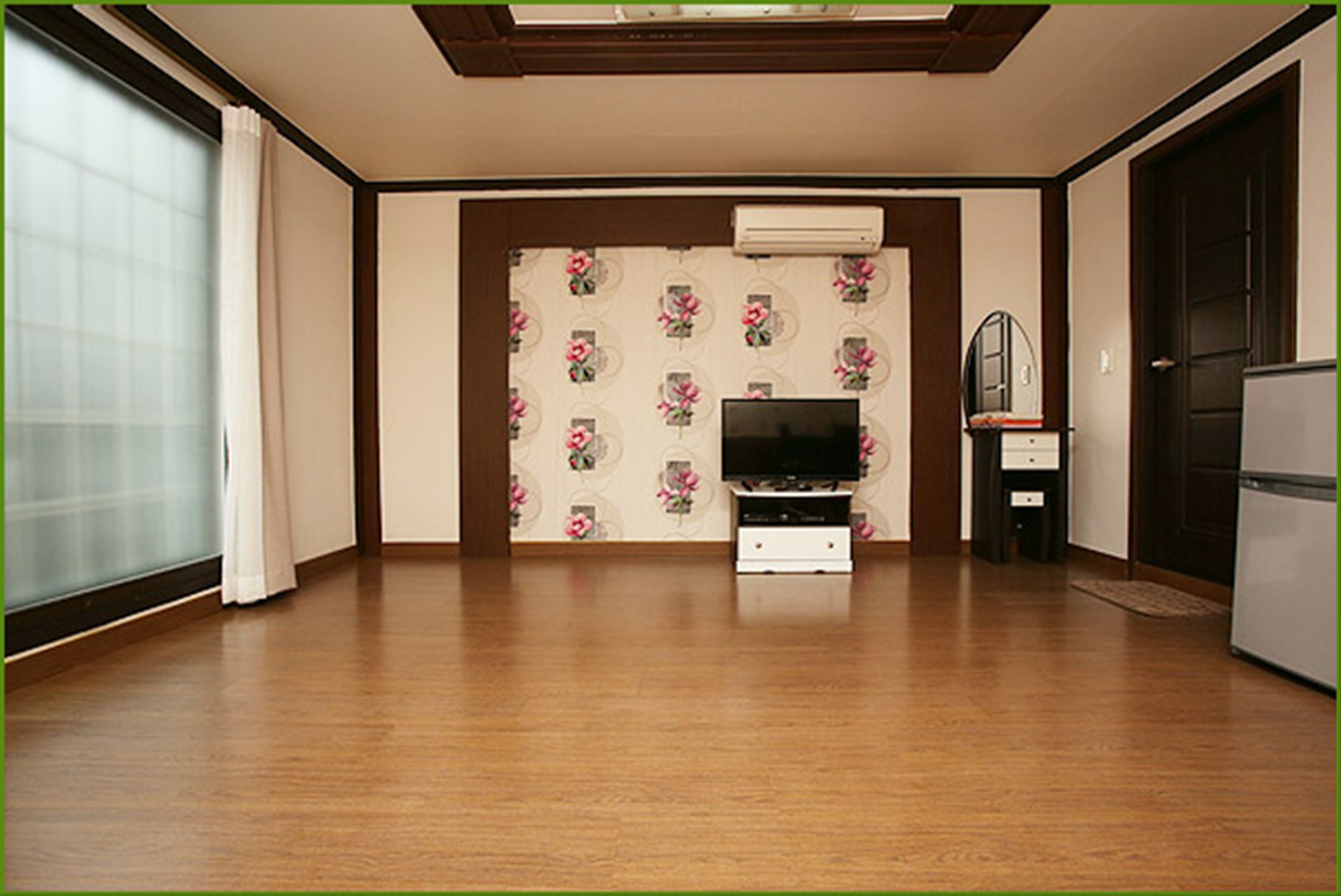 Una habitación con suelo de madera, un televisor sobre un mueble, un refrigerador y una puerta con ventana.