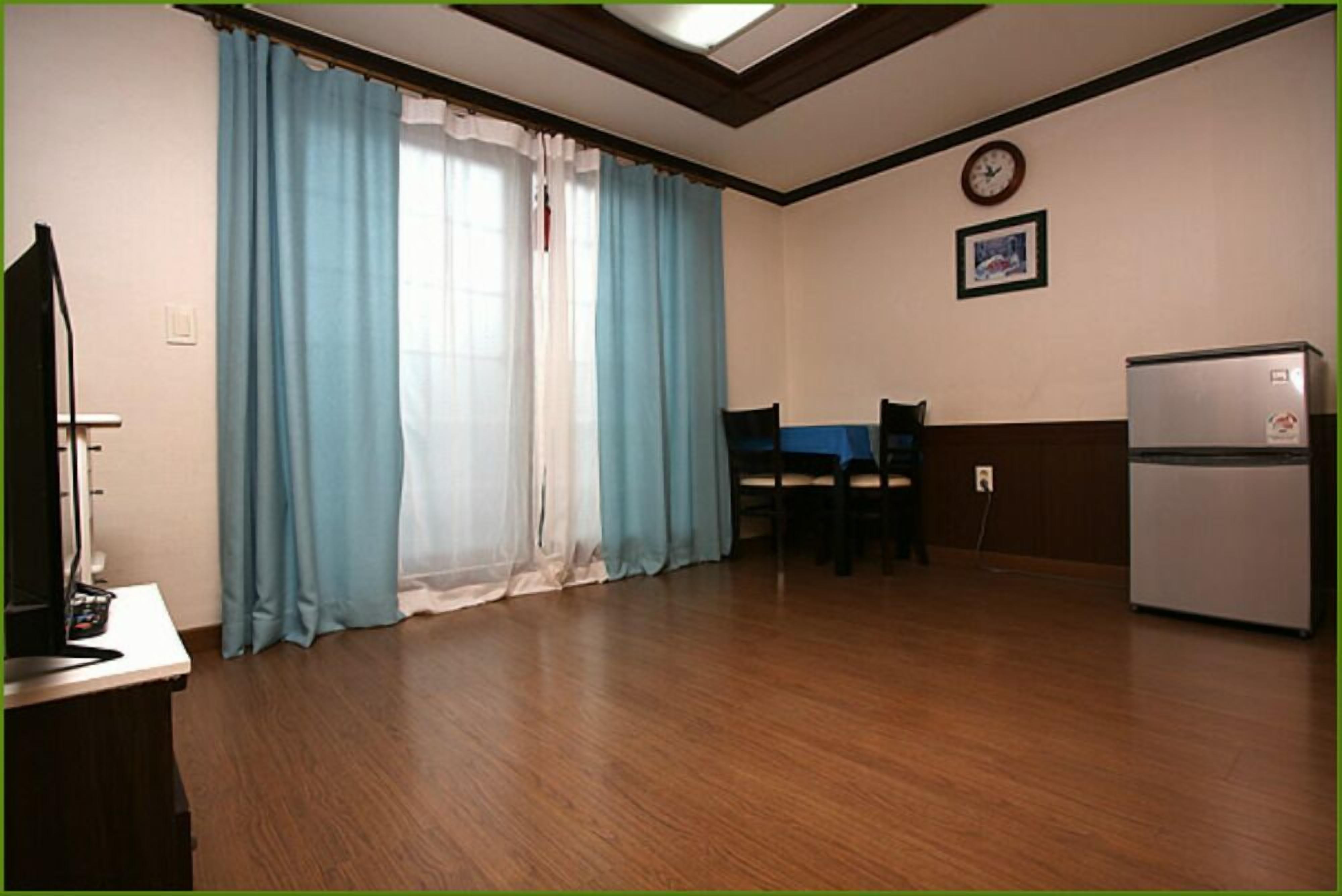 Una habitación con piso de madera, una mesa de comedor con sillas, un refrigerador, un televisor y una ventana con cortinas azules.