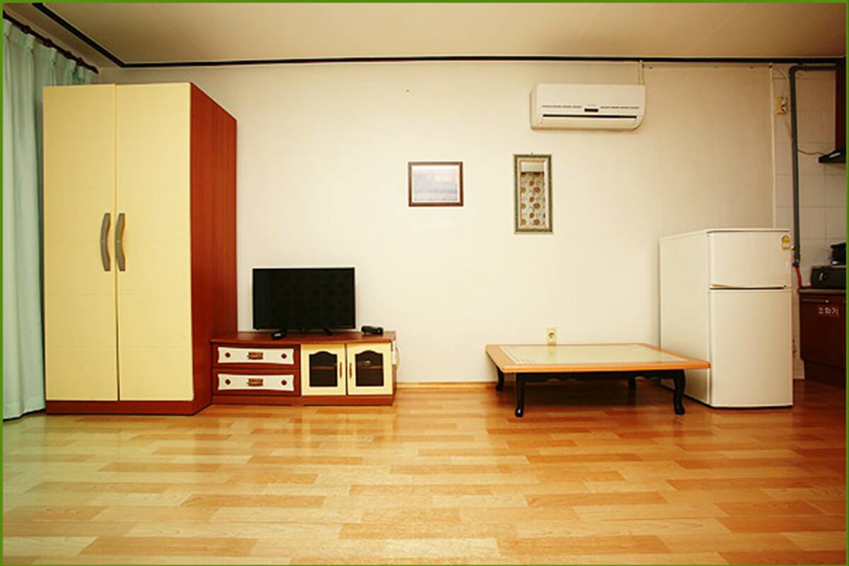 Une pièce avec un plancher en bois, un téléviseur posé sur un meuble, une petite table, un réfrigérateur blanc et une armoire.