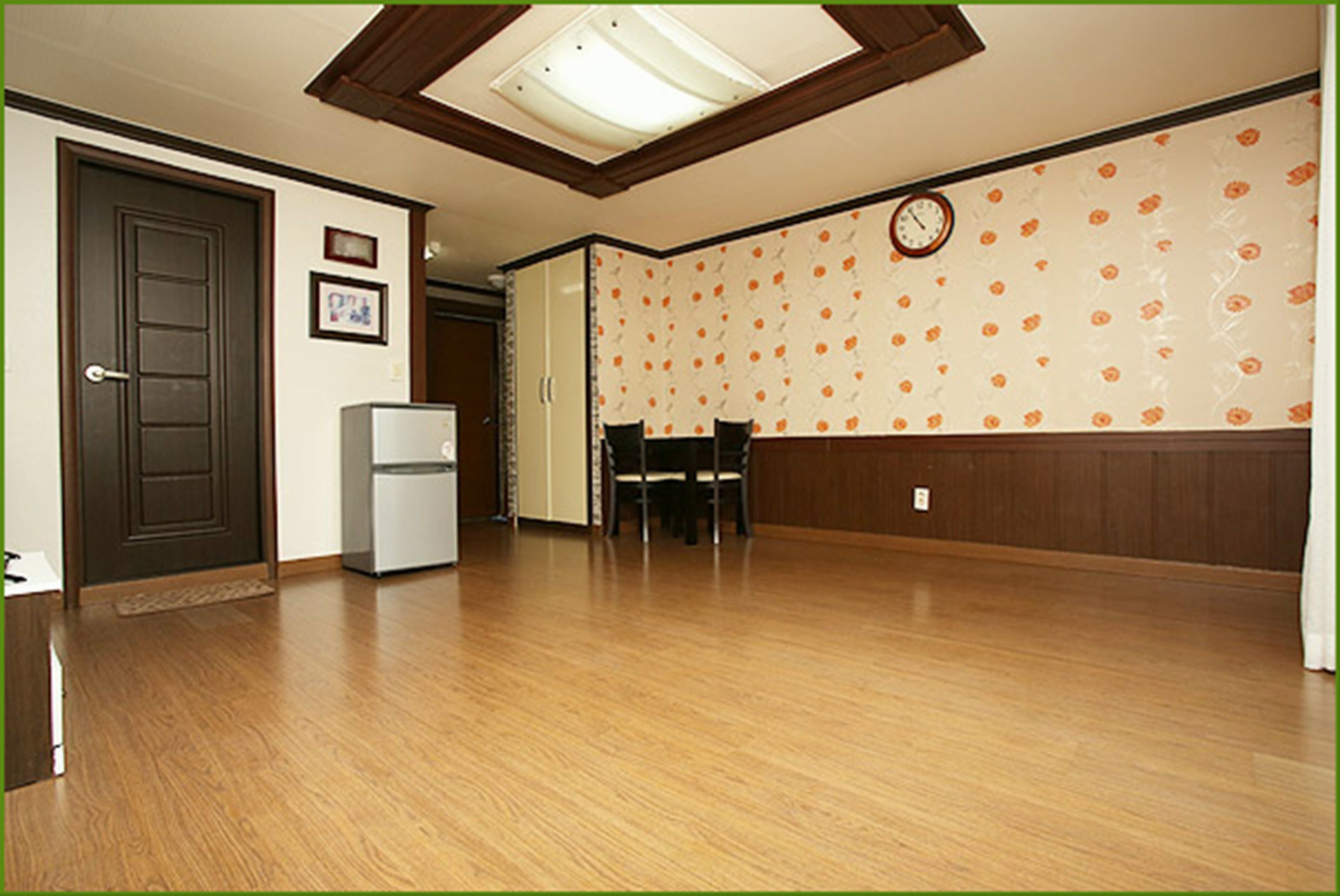 Ein Zimmer mit Holzboden, einer Tür, einem Kühlschrank, einem Esstisch mit Stühlen und einer Uhr an der Wand.