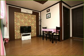 A room with a wooden floor, a television on a white stand, a dining table with chairs, and a door.