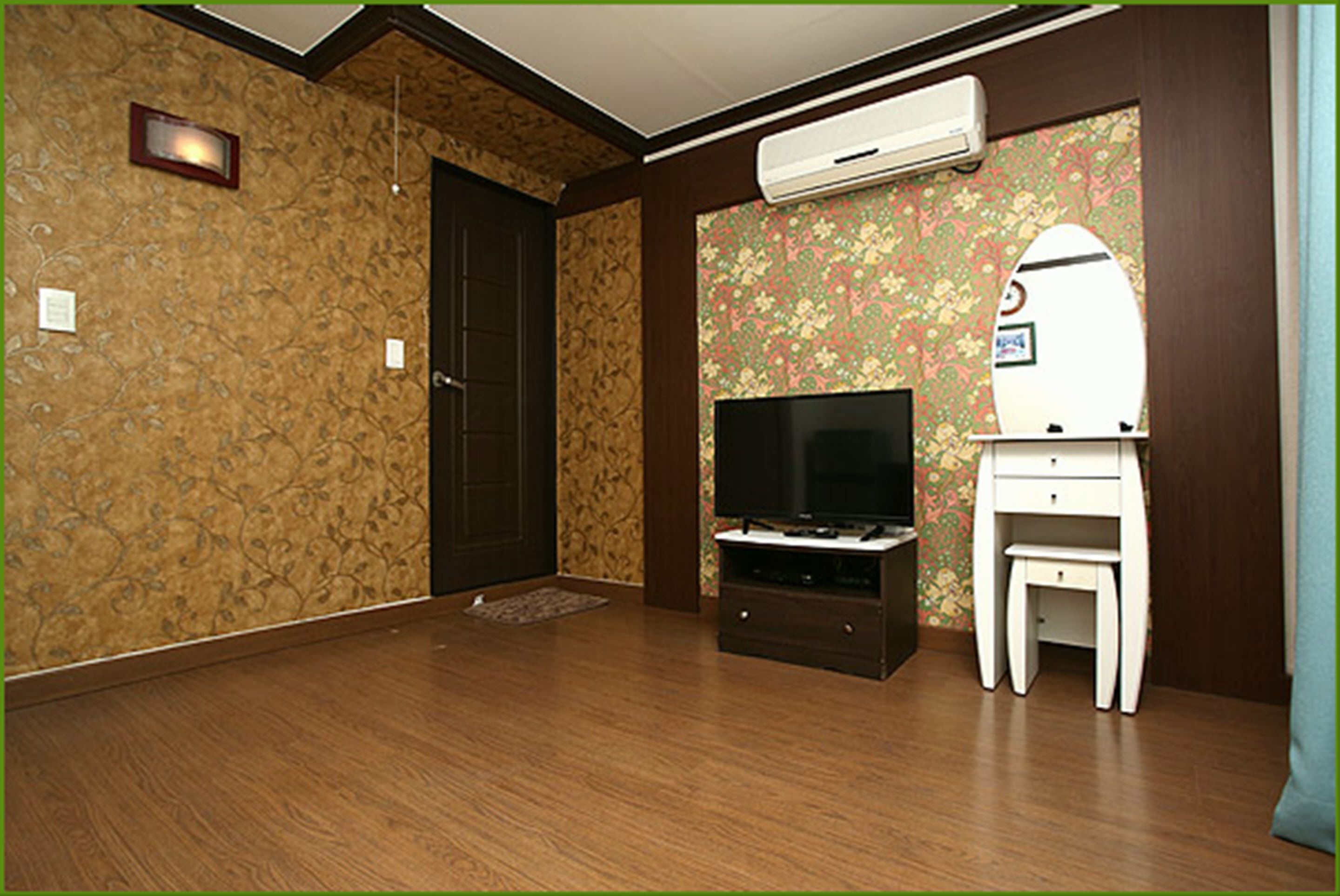 A room with wooden flooring, a dark door, a television on a stand, a small white table, and an air conditioning unit.