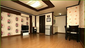 A room with wooden flooring, a flat-screen TV, a refrigerator, a dining table with chairs, and floral wallpaper.