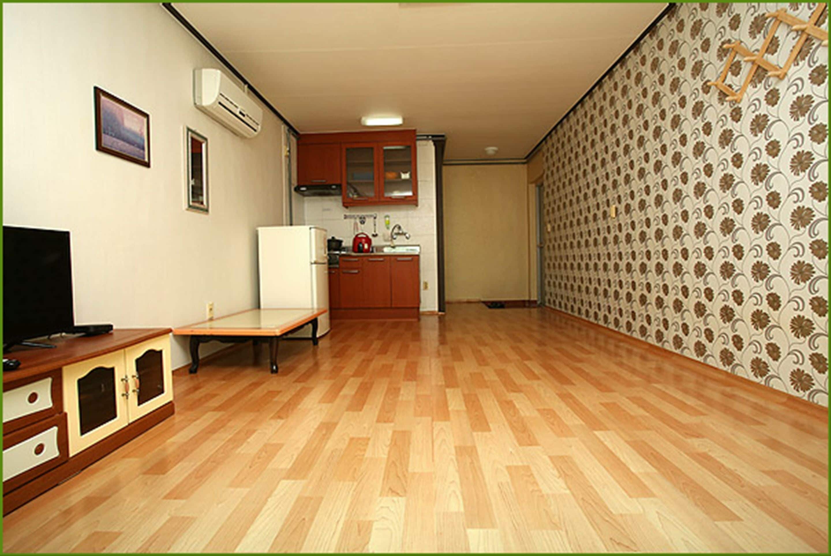 A room with a wooden floor, a television, a small table, a kitchen area with cabinets and a sink, and patterned wallpaper.