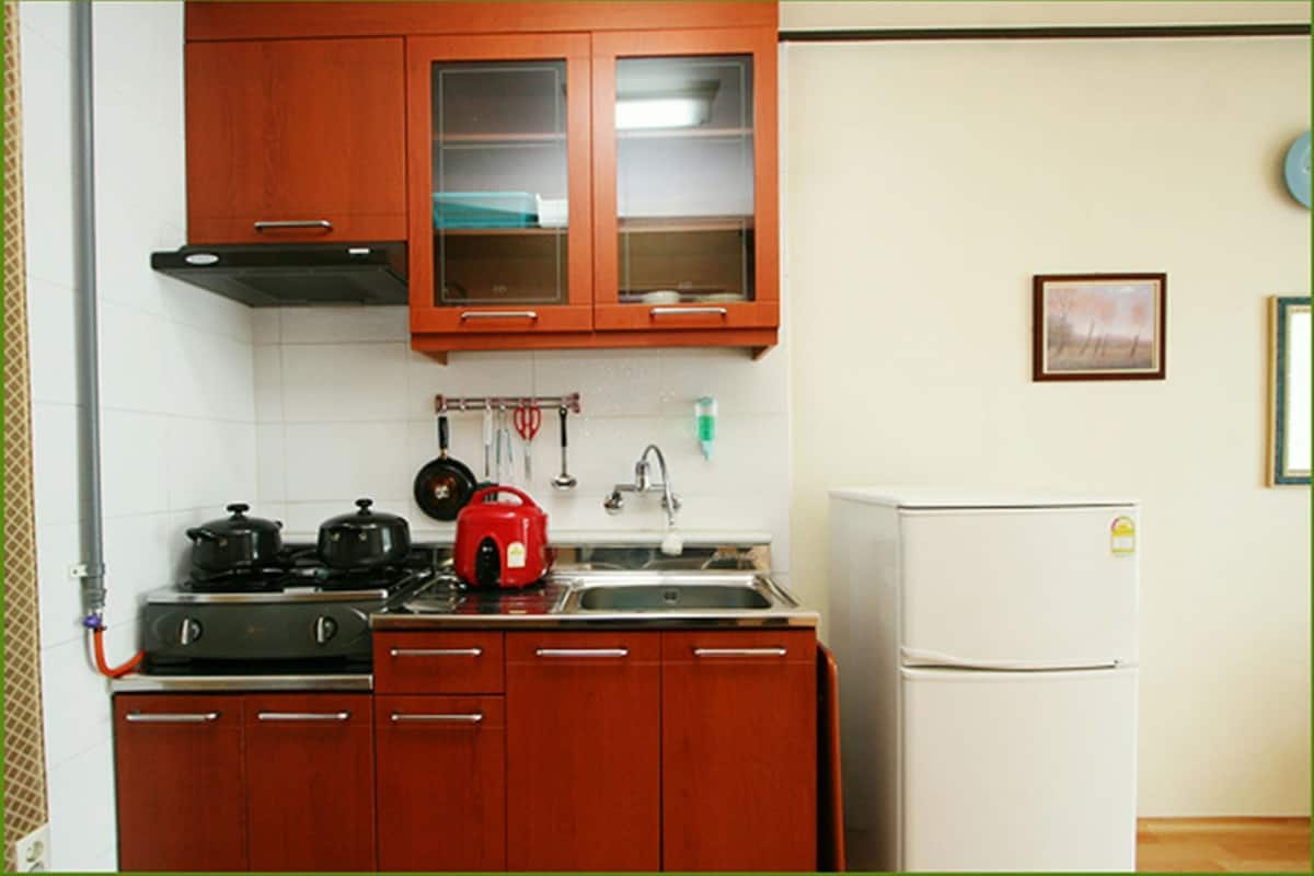 Une cuisine avec des meubles de rangement en bois, une cuisinière, une bouilloire rouge, un évier et un réfrigérateur blanc.