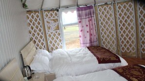 Ein Einzelbett mit weißer Bettwäsche und einer Decke in Gold und Rot, ein Nachttisch mit Wasserkocher und Tasse sowie ein Blick ins Freie durch ein Gitterfenster.