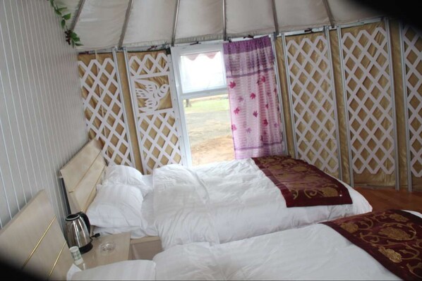 A single bed with white bedding and a red and gold throw blanket, a bedside table with a kettle and a cup, and a view of the outdoors through a lattice window.