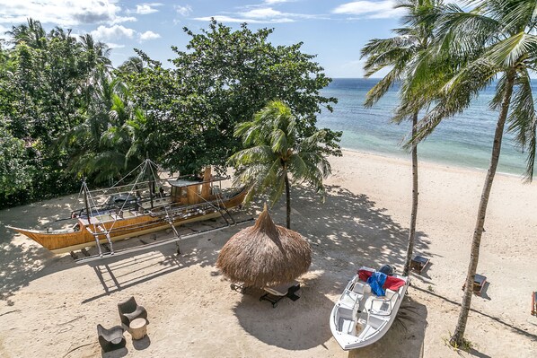 A beach with a thatched hut, a wooden boat, and a small boat docked near palm trees.