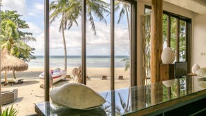 Vista a la playa desde una habitación con ventanas amplias, una mesa de vidrio y un paraguas de paja.