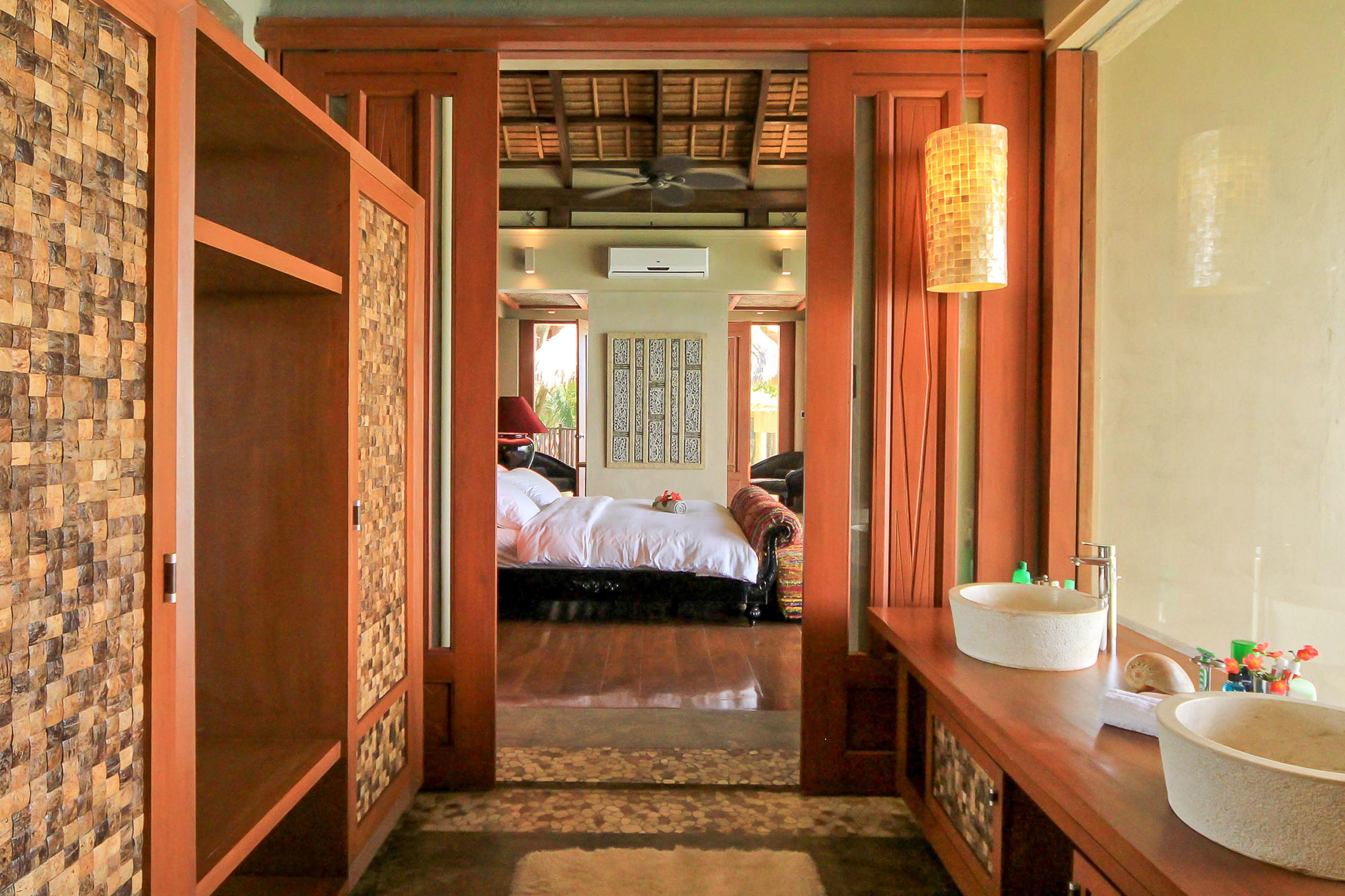 A bedroom with a bed, a wooden wardrobe, a bathroom with a large sink, and a ceiling fan.