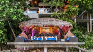 Un área de lounge al aire libre con techo de paja, asientos con cojines y una plataforma de piedra.