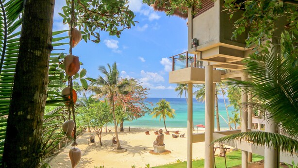 Vista de la playa con palmeras, arena y un edificio con balcones que dan al océano.