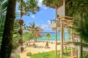 A beach view with palm trees, a sandy beach, and a building with balconies overlooking the ocean. - Den Pasar Nasog Villa (Malay)