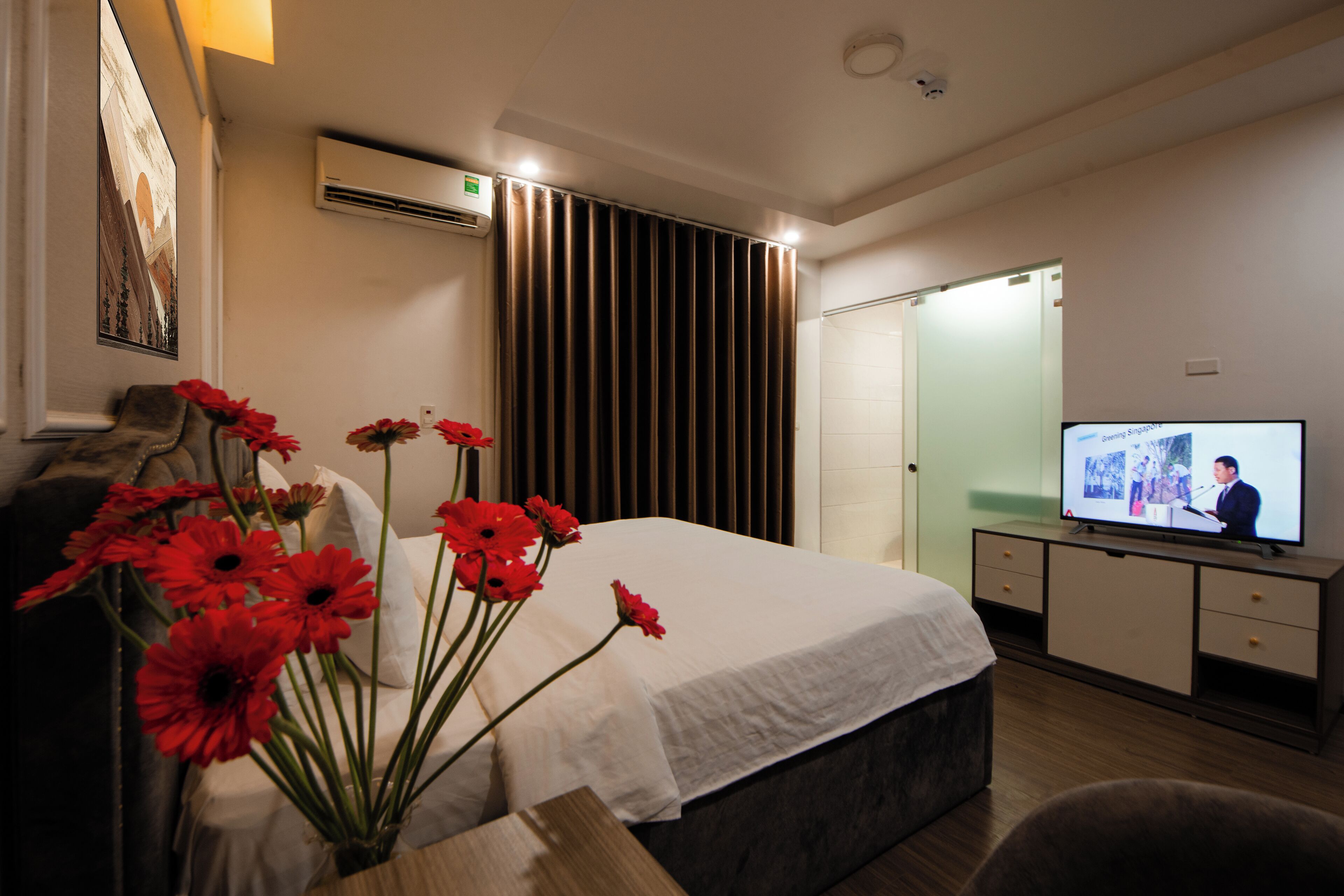 A hotel room with a bed, a television, a desk, and a vase of red flowers.