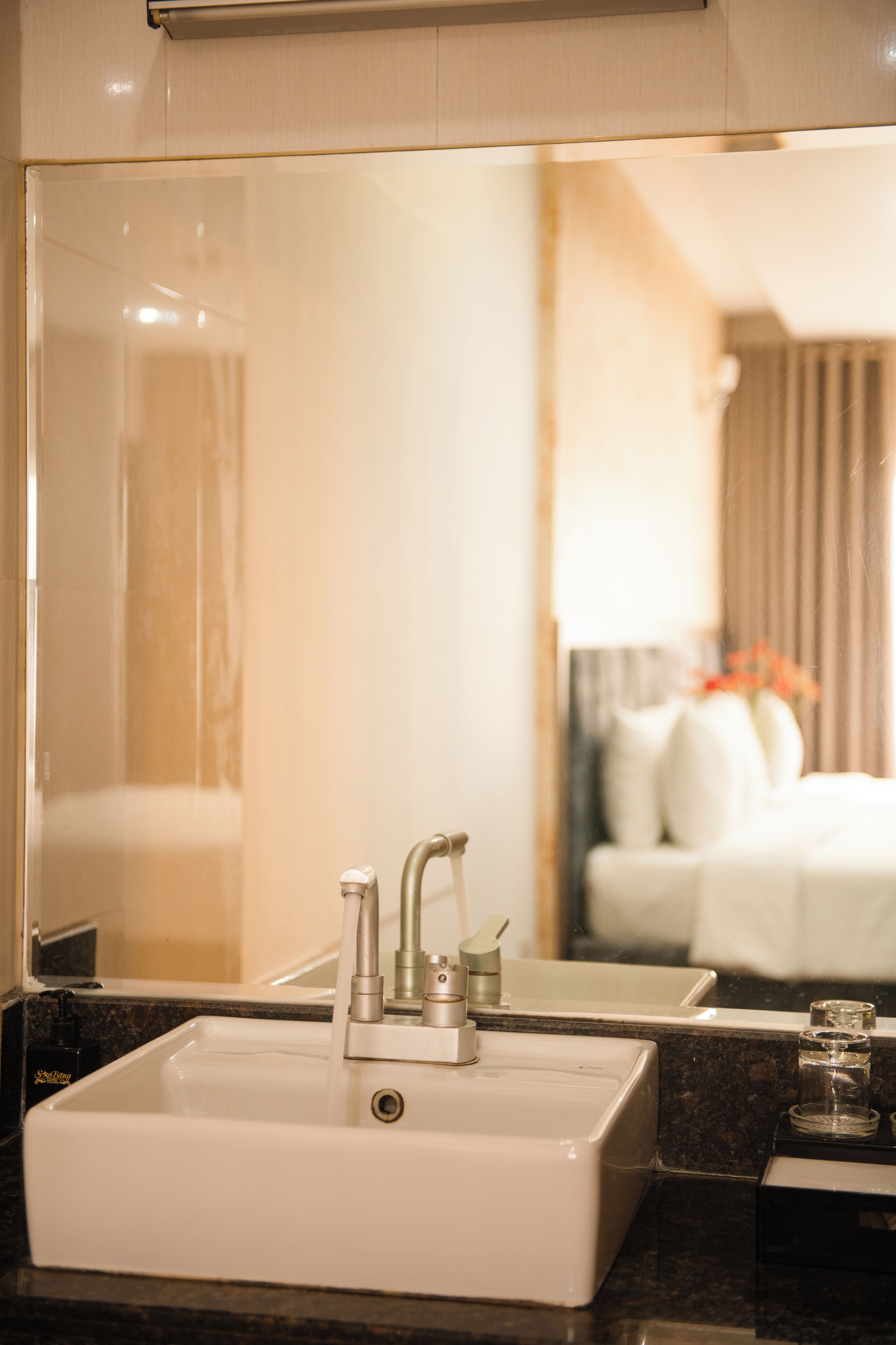 A bathroom with a white sink, a mirror, and a reflection of a bedroom with a bed and curtains.
