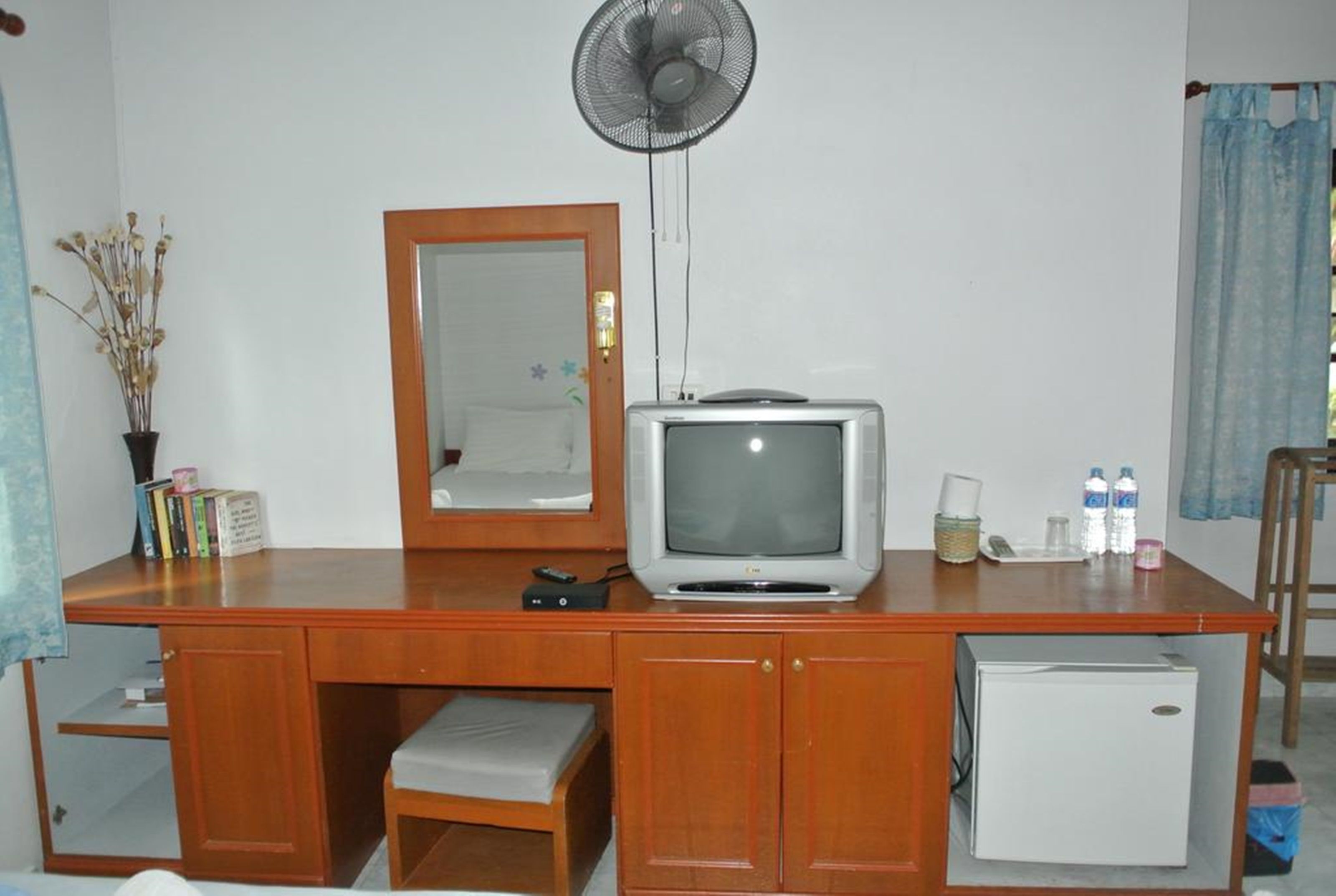 Una habitación compacta con escritorio de madera, refrigerador pequeño, televisor, espejo y ventilador de pie.