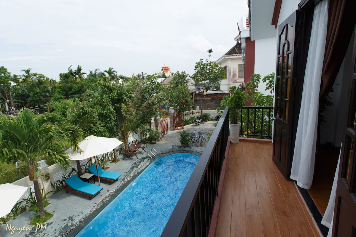 A swimming pool with lounge chairs and umbrellas, surrounded by greenery and a view of a traditional building in the background.