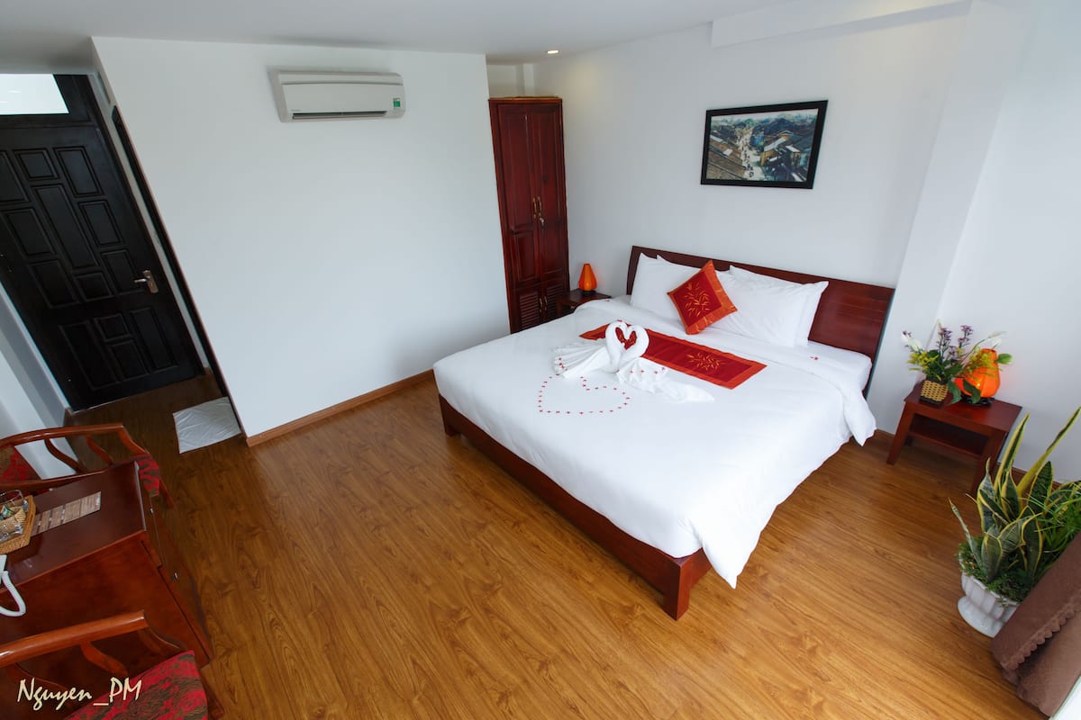 A hotel room with a wooden floor, a bed with white bedding and red accents, a wardrobe, a painting on the wall, and a small table with a plant.