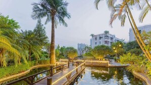 A wooden bridge over water, surrounded by palm trees and greenery, with a building in the background. - Delight Empire Hotel (Dongguan)