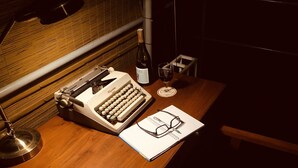 A wooden desk with a vintage typewriter, a wine bottle, a glass of wine, and a pair of glasses on a notebook. - Ahaspokuna by Eco Team (Balangoda)