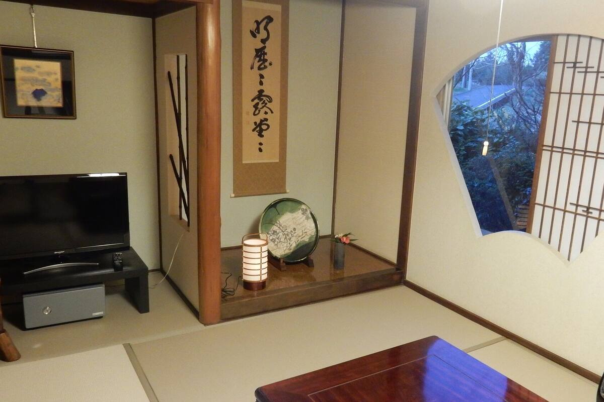 Ein traditioneller japanischer Raum mit Tatami-Böden, einem Fernseher, einem Shoji-Bildschirm und Blick auf einen Garten durch ein Fenster.