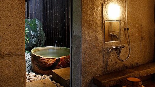 A stone-walled bathroom with a large, round, wooden bathtub and a wall-mounted shower. - Shouen (Izumo)