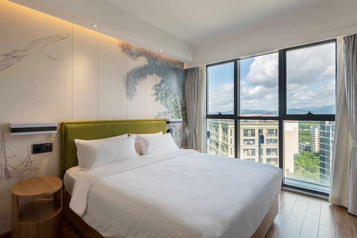 A modern hotel room with a large bed, a green headboard, and a view of the city through large windows.