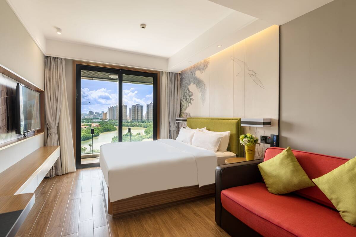 A hotel room with a large bed, a red sofa, a TV, and a city view through the window.