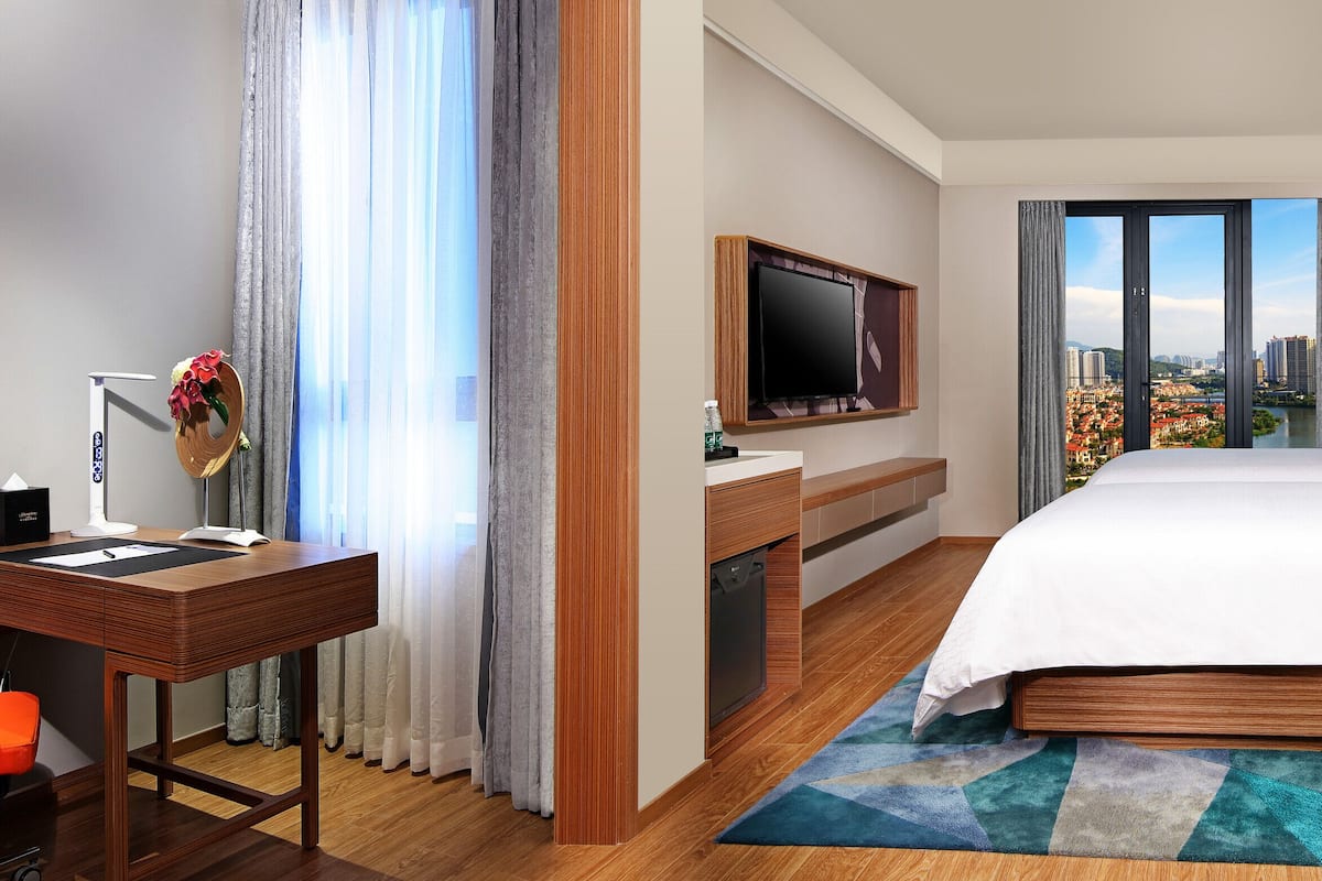 A hotel room with a bed, a desk with a chair, a television, and a view of a cityscape through large windows.