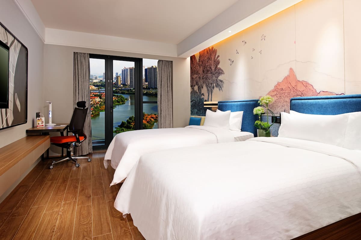 A hotel room with two beds, a desk with a chair, a TV, and a large window with a city view.