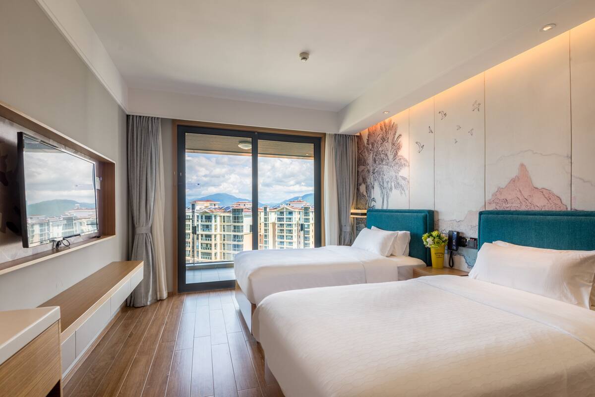 A hotel room with two beds, a TV, a desk, and a large window with a city view.