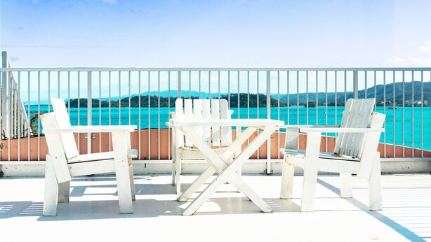 A deck with white chairs and a table, overlooking a turquoise sea and distant hills. - Amakara Okinawa (Nago)