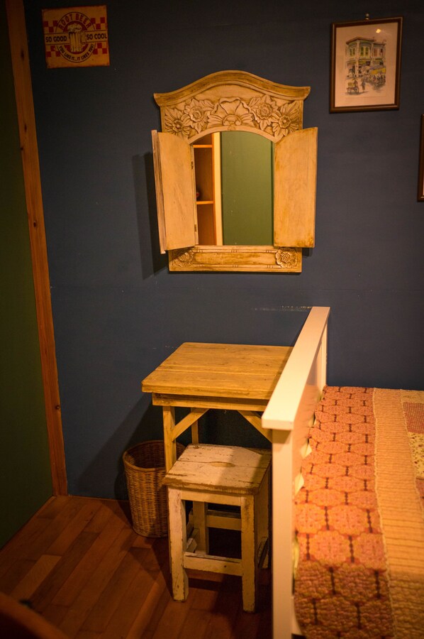 Una habitación pequeña con una mesa de madera, un taburete, un espejo con un gabinete abierto, una cesta de mimbre y una alfombra estampada.