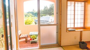 A room with a sliding glass door leading to a balcony, a small table with a bench, and a window with a view of greenery and a building. - Amakara Okinawa (Nago)