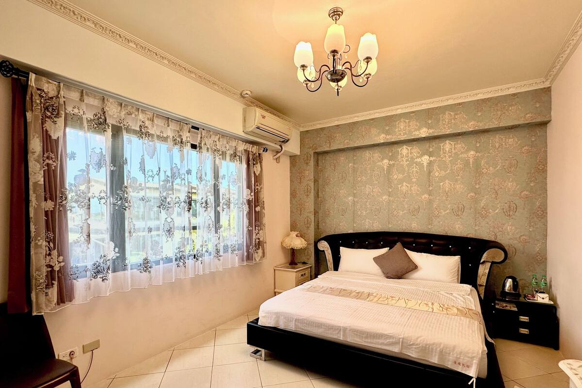 Un dormitorio con cama, mesita de noche, una silla y una ventana con cortinas.