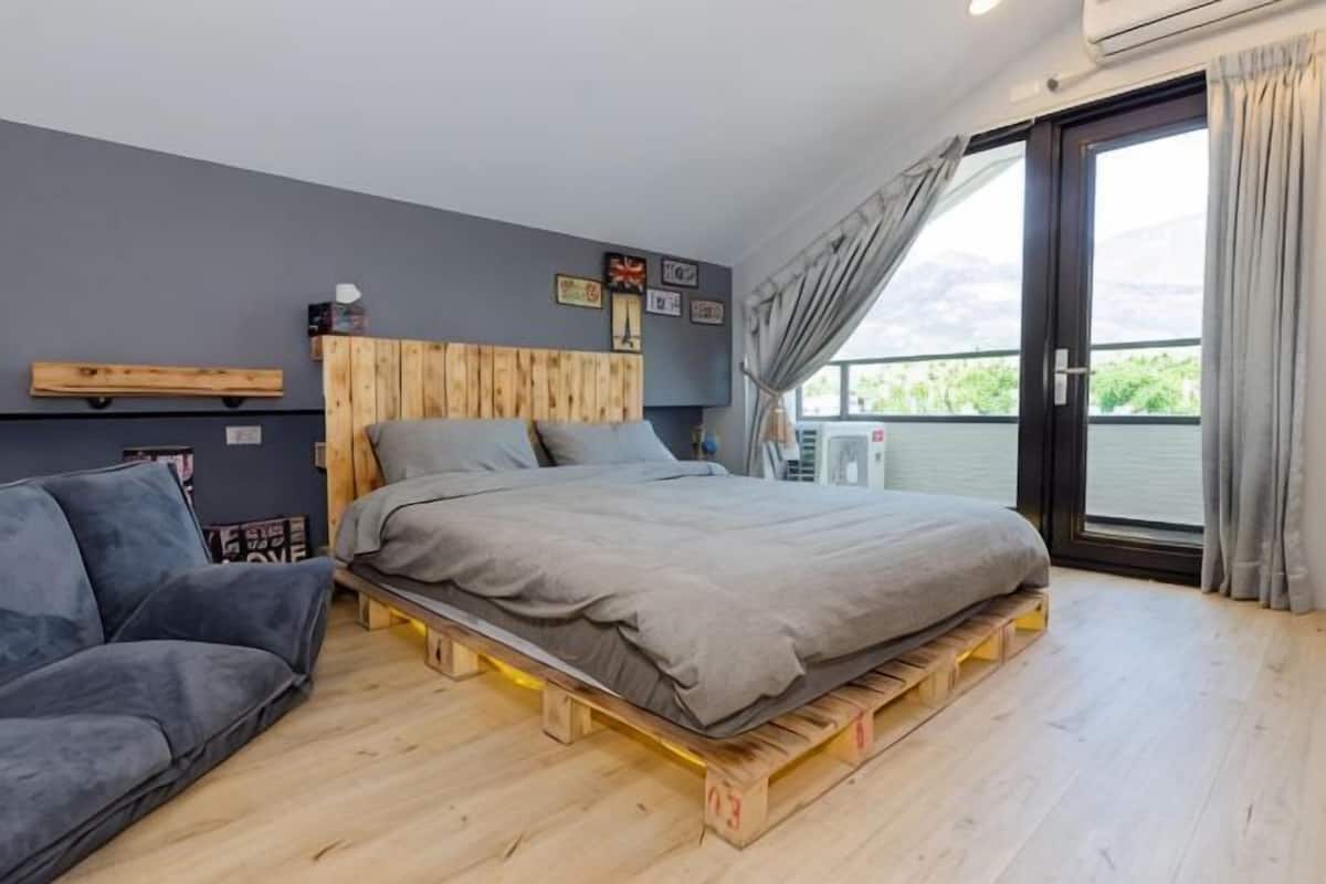 Un dormitorio con cama sobre plataforma de madera, un sofá, una ventana con cortinas y vistas al exterior.